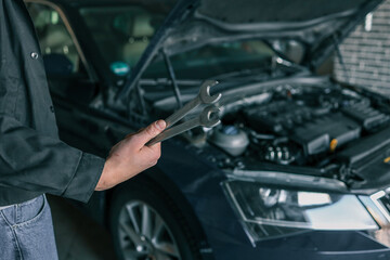 Poster - A mechanic holding wrenches in front of an open car hood in a garage. The image emphasizes automotive repair tools and professional car maintenance in a workshop setting