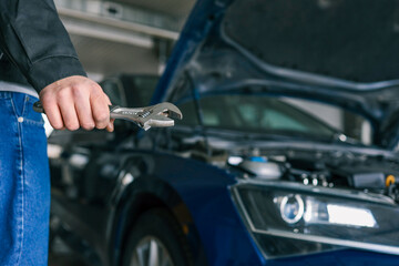 Fototapete - Hand holding an adjustable wrench in an automotive repair shop. A blue car with an open hood is visible in the background, highlighting repair or maintenance activity