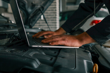 Fototapete - Close-up of a mechanic's hands typing on a laptop placed on a car engine in a workshop, showcasing the integration of technology in vehicle diagnostics and repairs