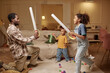 © Seventyfour - Side view of African American father playing with children fighting with toy swords, while reciting lines of noble knight standing on one knee in messy living room