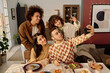 © AnnaStills - High angle shot of biracial company of friends sitting at festive table and taking selfie together