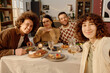 © AnnaStills - Medium close up of biracial friends sitting at served festive table looking at camera and posing