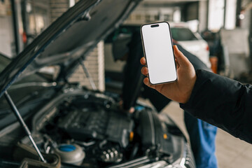 Fototapete - Hand holding a smartphone with a blank screen in front of an open car hood in a workshop setting, with blurred mechanics and tools in the background