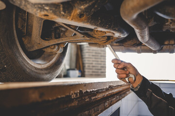 Aufkleber - Mechanic's hand holding a wrench, working underneath a car on a lift. The view shows a close-up of the suspension and exhaust system in a garage setting