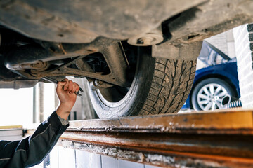 Aufkleber - Mechanic's hand holding a wrench, working underneath a car on a lift. The view shows a close-up of the suspension and exhaust system in a garage setting