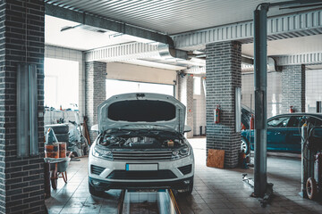Fototapete - Interior view of an empty automotive repair shop with a car on a lift, its hood open. The workshop features tiled floors, brick walls, and vehicle service equipment in the background