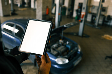 Fototapete - Mechanic holding a tablet with a blank screen in a car repair shop. A blue car with an open hood is in the background, and the workshop has visible car lifts and tools