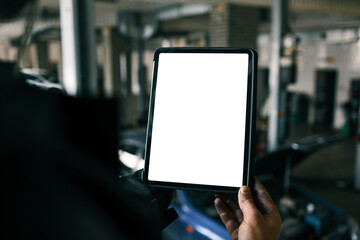 Fototapete - Mechanic holding a tablet with a blank screen in a car repair shop. A blue car with an open hood is in the background, and the workshop has visible car lifts and tools