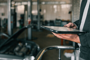 Fototapete - Mechanic holding a tablet in an automotive workshop, inspecting car diagnostics and maintenance information, with a blurred repair area in the background