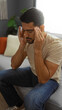 © Krakenimages.com - Young man with beard sitting on couch in living room with hands on temples looking stressed indoors at home with natural light from window