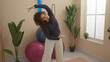 © Krakenimages.com - Young woman exercising indoors in a gym with plants and exercise balls, performing a side stretch with arms extended.