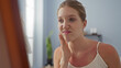 © Krakenimages.com - Young woman with blonde hair in a spa room examining her skin near a mirror, displaying a displeased expression.