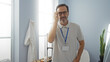 © Krakenimages.com - Middle-aged hispanic man inside a clinic room, wearing glasses and a name badge, standing confidently indoors in a professional setting.