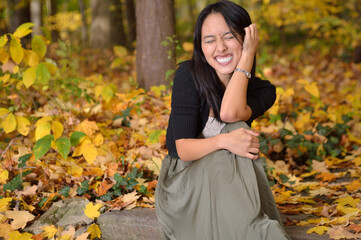 A beautiful young Asian woman in a dress and black sweater sits among bright yellow leaves - autumn - grinning