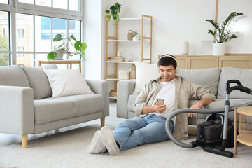 Wall Mural - Young man with mobile phone and vacuum cleaner listening to music at home