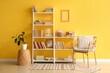 © Pixel-Shot - Shelving unit with books, armchair and houseplant near yellow wall in room