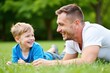 © SimpleDesignStudio - Heartwarming Bond Between Father and Son Lying on Green Grass in Natural Park Setting, Joyful Expressions in a Beautiful Daylight Environment