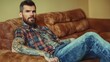 © JO BLA CO - A man with tattoos and a beard is sitting on a couch. He is wearing a plaid shirt and jeans