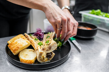 Wall Mural - A chef carefully garnishes a plate of bone marrow with fresh herbs and condiments, accompanied by crispy toasted bread. A vibrant kitchen setting enhances the culinary atmosphere.