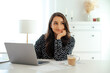 © Kateryna - Thoughtful pretty woman sitting at a desk with a laptop and a cup of coffee, resting her head on her hand, looking away and smiling, showing a relaxed and optimistic mood in a bright modern office