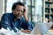 © Наталья Лазарева - Thoughtful African American man with glasses sitting at desk surrounded by crumpled papers and using laptop,representing workplace challenges,problem-solving,determination in modern office environment