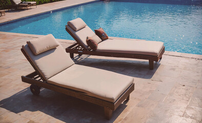 Poster - Two lounge chairs are placed on a tiled pool deck. The chairs are tan and have pillows on them. The pool is surrounded by a tiled area, and there are several chairs and a bench nearby. The scene is