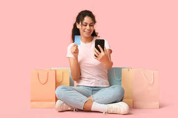 Wall Mural - Young woman sitting with credit card, shopping bags and mobile phone on pink background