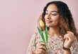 © Sandor - Hispanic woman with curly hair holding and smelling a yellow tulip with a pink background