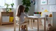 © Jaroon - A little girl is sitting at a wooden table with a box of crayons in front of her. She is focused on her drawing, possibly a picture of a tree or a flower. The room is decorated with various items