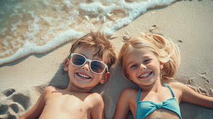 A close-up shot of the happy little boy and girl with blond hair and sunglasses lying on the sandy beach on a sunny day with waves in the background. Summer vacation concept.