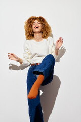 Poster - Happy woman with curly hair laughing joyfully in a white room, wearing a light sweater and blue jeans, showcasing a carefree attitude and vibrant energy