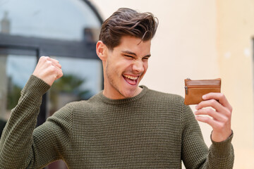 Wall Mural - Young caucasian man holding a wallet at outdoors celebrating a victory