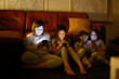 © natalialeb - Technology and childhood: four kids seated by the sofa, glued to their phones, authentic dark style.