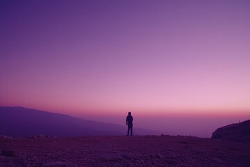 Fototapete - A solitary figure stands on a rocky hilltop, silhouetted against a twilight sky while gazing into the distant horizon, evoking a sense of mystery