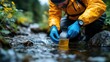 © PICK YOUR POV - A person in a yellow jacket collects water from a stream using a bottle for testing or analysis.