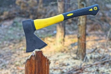 one small sharp iron black dangerous modern plastic yellow handle industrial hatchet sticking out of a wooden stump during the day in the forest