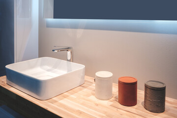 A white sink sits on a wooden countertop with three containers next to it. The sink is rectangular and has a chrome faucet. The containers are of different sizes and colors