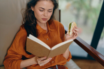 Canvas Print - A young woman enjoying a sandwich while reading a book, dressed in an orange shirt, showcasing a relaxed atmosphere, perfect for lifestyle and wellness themes