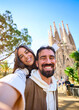 © CarlosBarquero - Vertical. Young mixed race couple taking a selfie in front of Sagrada Familia in Barcelona. Smiling Husband and wife on street of city enjoying vacation happy and smiling. Funny tourism people
