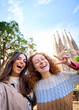 © CarlosBarquero - Vertical. Young funny caucasian women friends taking selfie in front of Sagrada Familia in Barcelona. Smiling beautiful girl on street of European city enjoying vacation happy. Tourism people