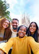 © CarlosBarquero - Vertical. Group diverse funny young tourist friends posing piggyback with hands joined in air taking selfie together in front of Sagrada Familia in Barcelona. Happy people enjoy holidays outdoor