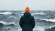 © Sirichai - Facing the Storm:  A solitary figure, wearing a bright orange beanie, stands stoic and resolute against the backdrop of a tempestuous sea.