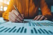 © Jakrapan - A person writes notes and analyzes data on printed graphs and charts, with soft lighting illuminating the workspace.