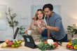 © crizzystudio - Young asian couple having a video call showing their hands while preparing ingredients for cooking in a kitchen at home, vegetables and fruits on table