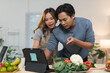 © crizzystudio - Young couple wearing aprons following online cooking course using tablet and preparing healthy vegetable salad for lunch in kitchen at home