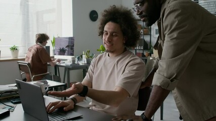 Positive young programmer showing new software to coworker while both men looking at laptop screen in office