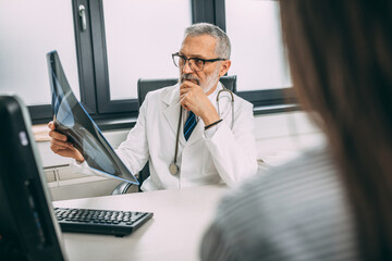 A mature radiologist in a white coat is seriously looking at an x-ray of a sick female patient while sitting in the doctor's office.