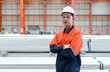 © chadchai - A worker in an orange jacket, hardhat, protective glove and safety glass stand confidently with arm crossed in front of a pile of material covered in plastic on the floor.