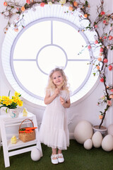 Poster - A little curly blonde hair girl in white dress with with large Easter eggs in spring interior decoration room. Happy smiling child girl with chick and carrot in basket near decorated floral window	
