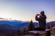 © maryviolet - Man taking picture of mountain landscape using smartphone at sunset sitting on bench enjoying view.